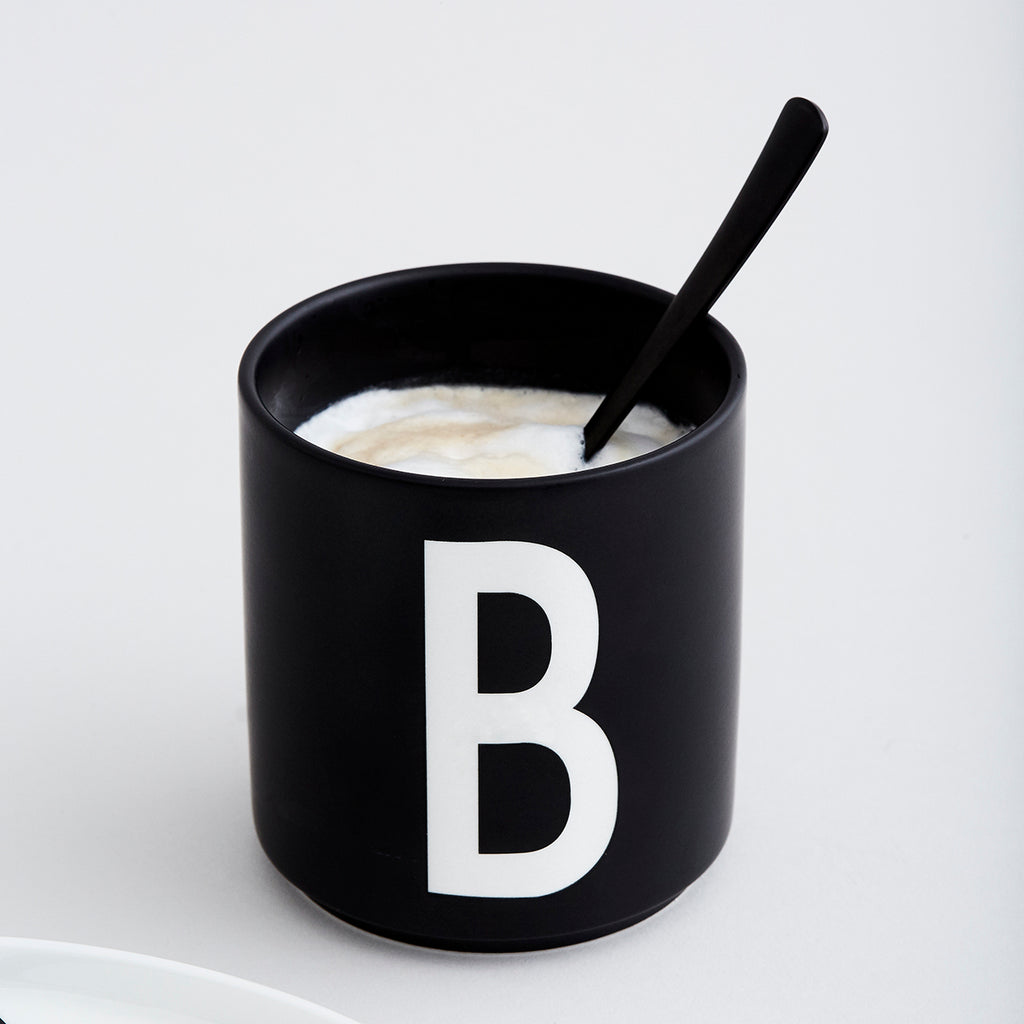 Schwarze Tasse mit weißem B, mit Kaffee und Löffel