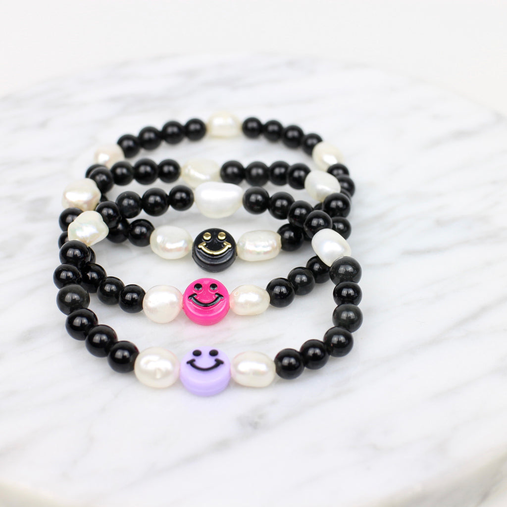 3 Black Smiley Armbänder. Schwarze Perlen, 5 weiße Süßwasserperlen und ein Smiley, bei einem Armband fliederfarbener Smiley, bei dem zweiten pinkfarbener Smiley, beim dritten ein schwarzer Smiley