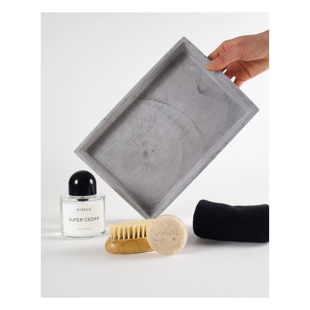 Eine Hand hält das graue Tablett aus Beton frontal zur Kamera. Auf einen Tisch sind Bürste, Seife und Handtuch dekoriert