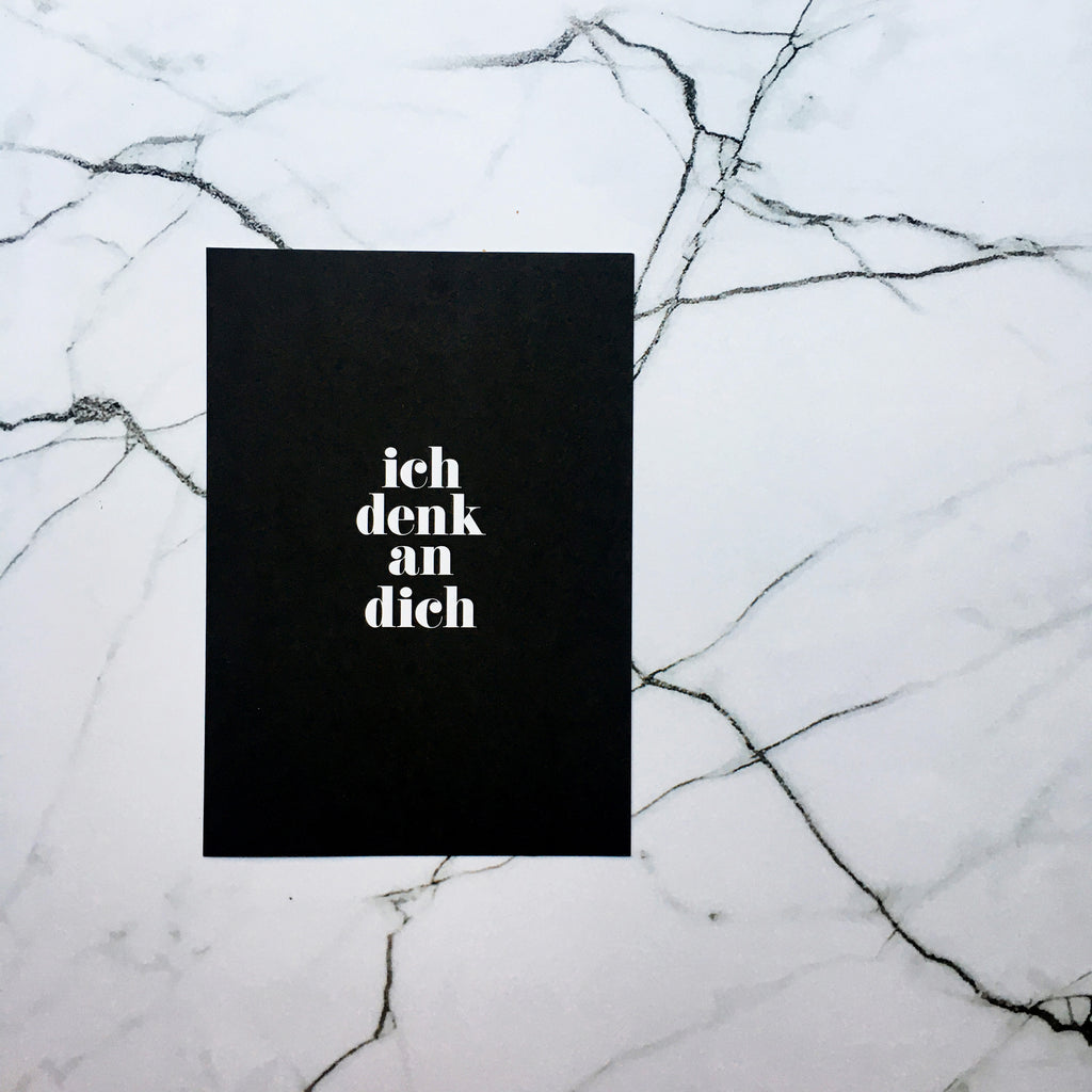 schwarze Postkarte mit weißer Schrift "ich denk an dich" vor marmoriertem Hintergrund