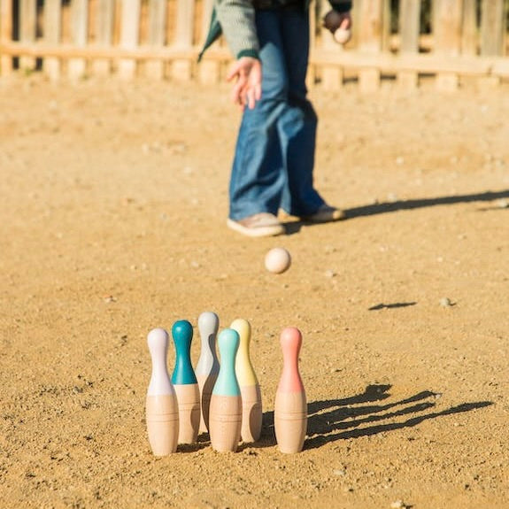 Bowling Pins X Set stehen draußen auf einem Sandboden und ein Model wirft eine Holzkugel, um sie umzuwerfen