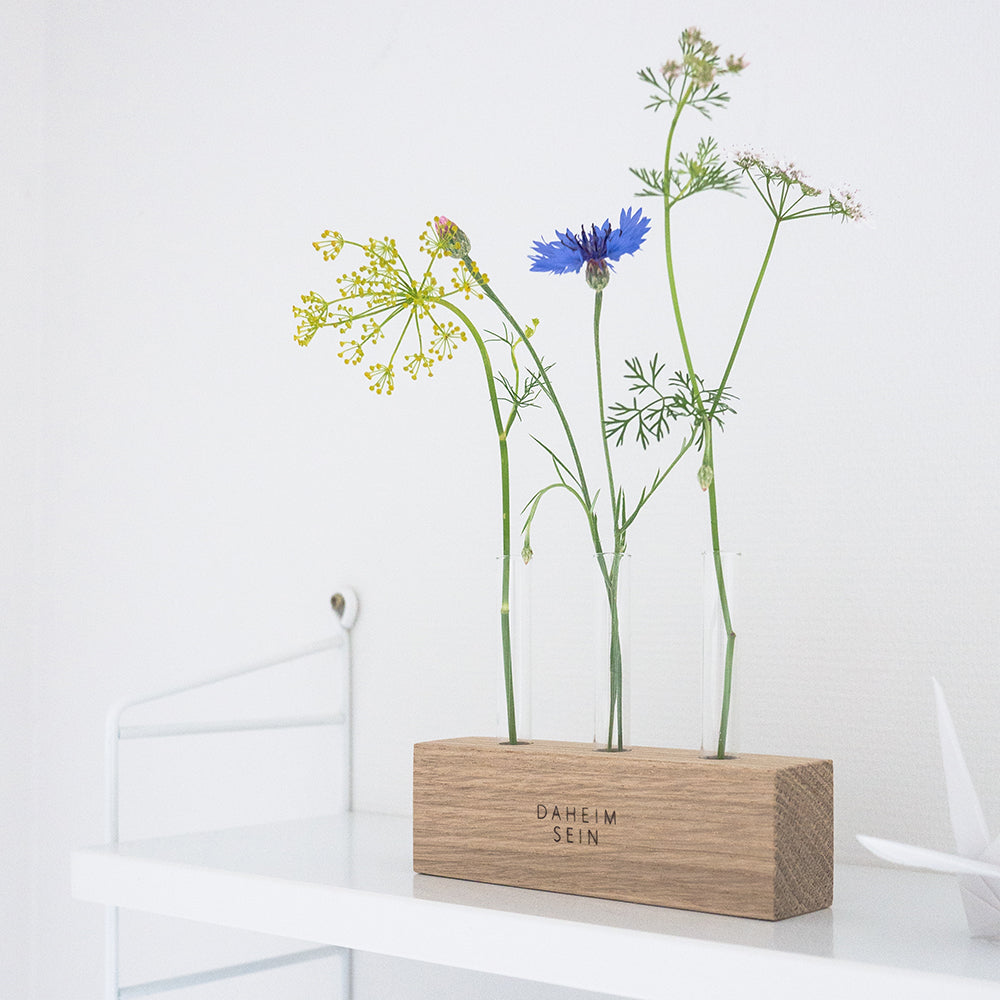 Holzleiste mit Aufschrift "daheim sein" mit drei Reagenzgläsern als kleine Blumenvasen, mit Blumen dekoriert