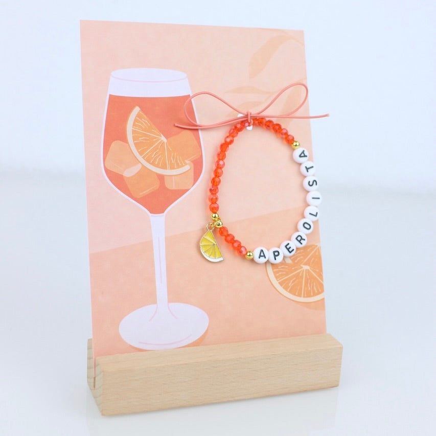 Armband mit orangenen Glasperlen, einem Anhänger in Form einer halben Orangenscheibe und Buchstaben "Aperolista" schwarz auf weiß, dekoriert an einer orangenen Postkarte mit Aperolglas