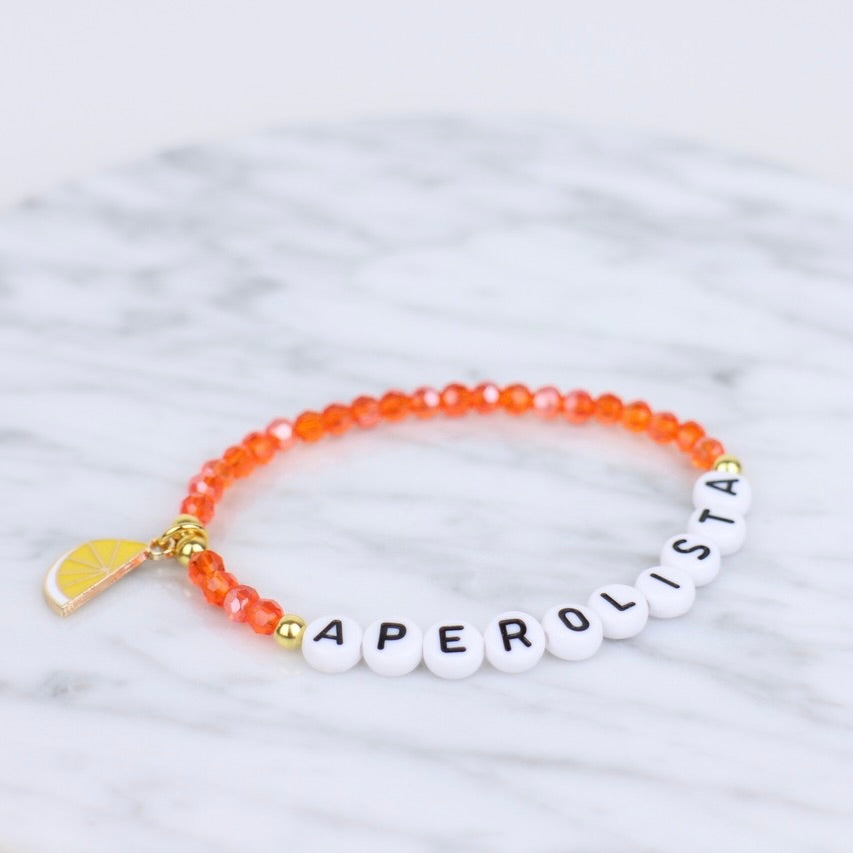 Armband mit orangenen Glasperlen, einem Anhänger in Form einer halben Orangenscheibe und Buchstaben "Aperolista" schwarz auf weiß
