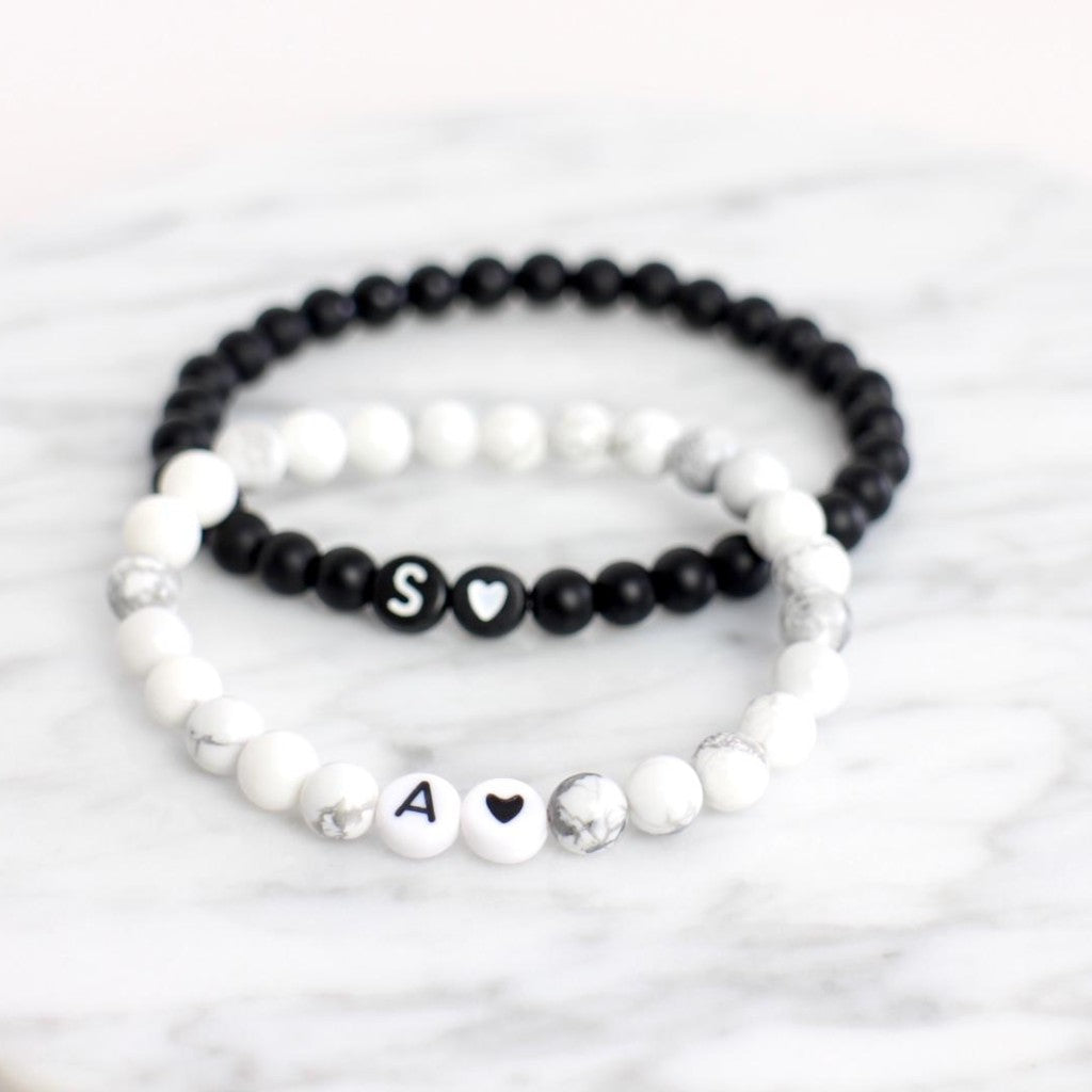 Ein weißes Armband mit schwarzem Buchstaben "A" und schwarzem Herz. Ein schwarzes Armband mit weißem Buchstaben "S" und weißem Herz.