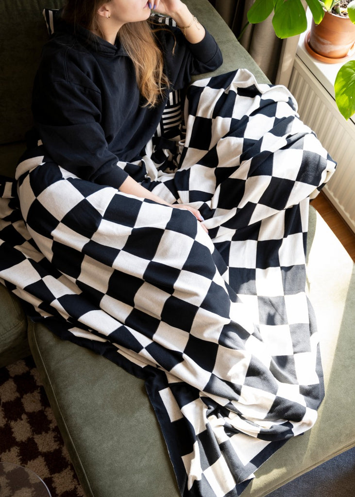 Ein Model liegt auf einer grünen Couch, zugedeckt mit der Ecru-Schwarz X Caro Plaid Kuscheldecke