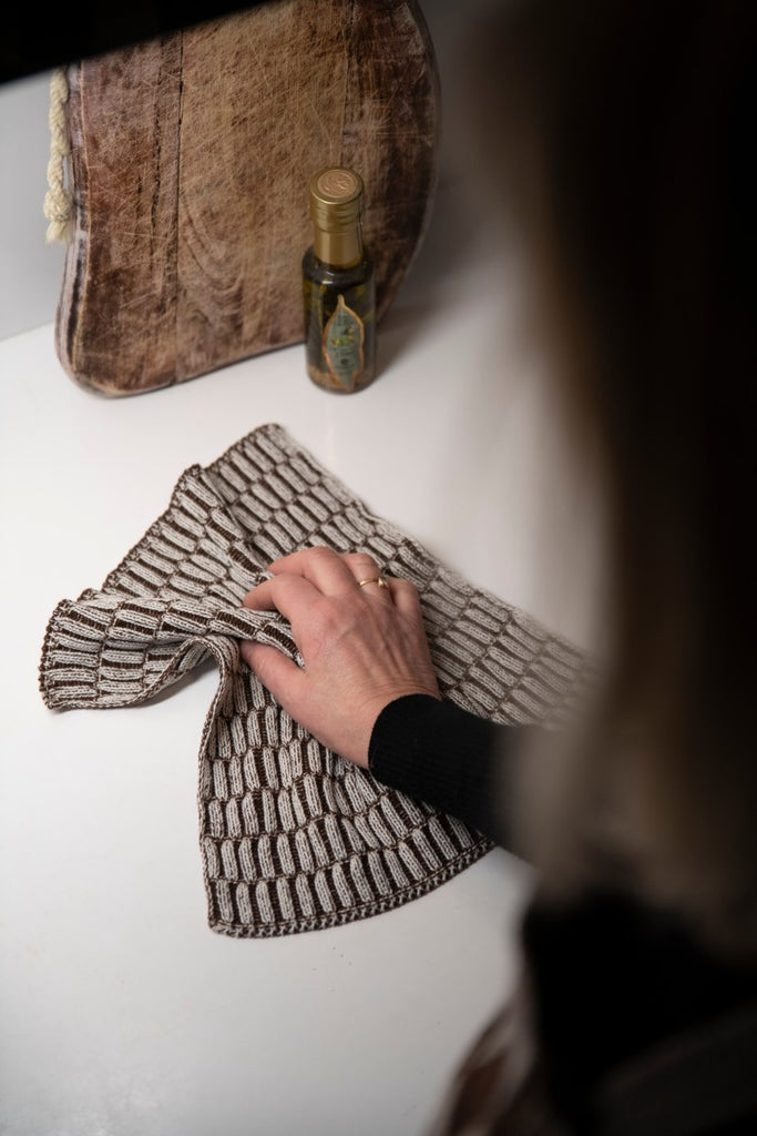 Es wird mit einem gestrickten Spültuch in Brauntönen der Tisch abgewischt