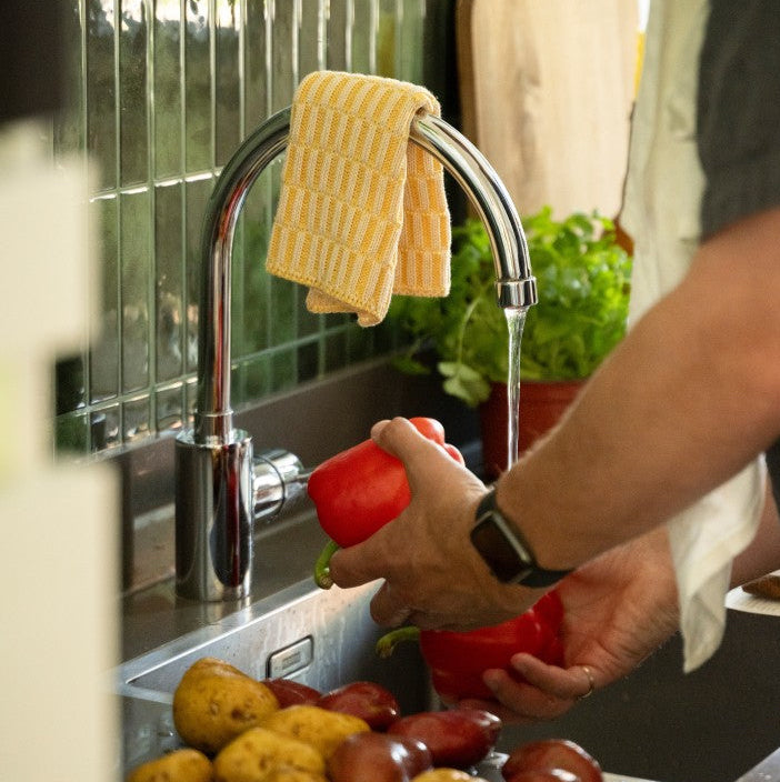 Ecru-Goldgelb X Gestricktes Spültuch, abgelegt auf einem Wasserhahn in der Küche, es wird Gemüse gewaschen