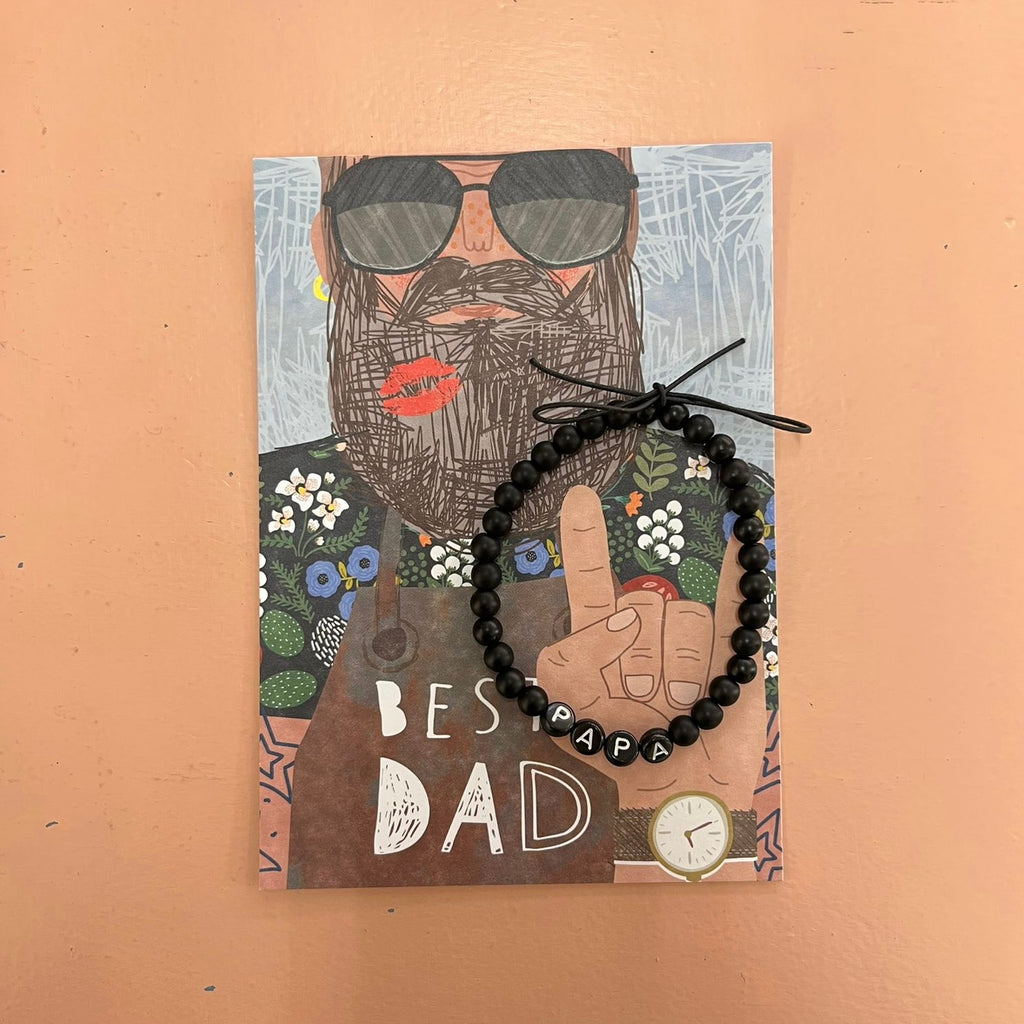 Best Dad X Armband Papa, schwarze Perlen und weiße Buchstaben "Papa" auf schwarzen Perlen. Befestigt an einer Postkarte mit einem gezeichneten Mann mit Vollbart und Sonnenbrille mit Text "Best Dad"
